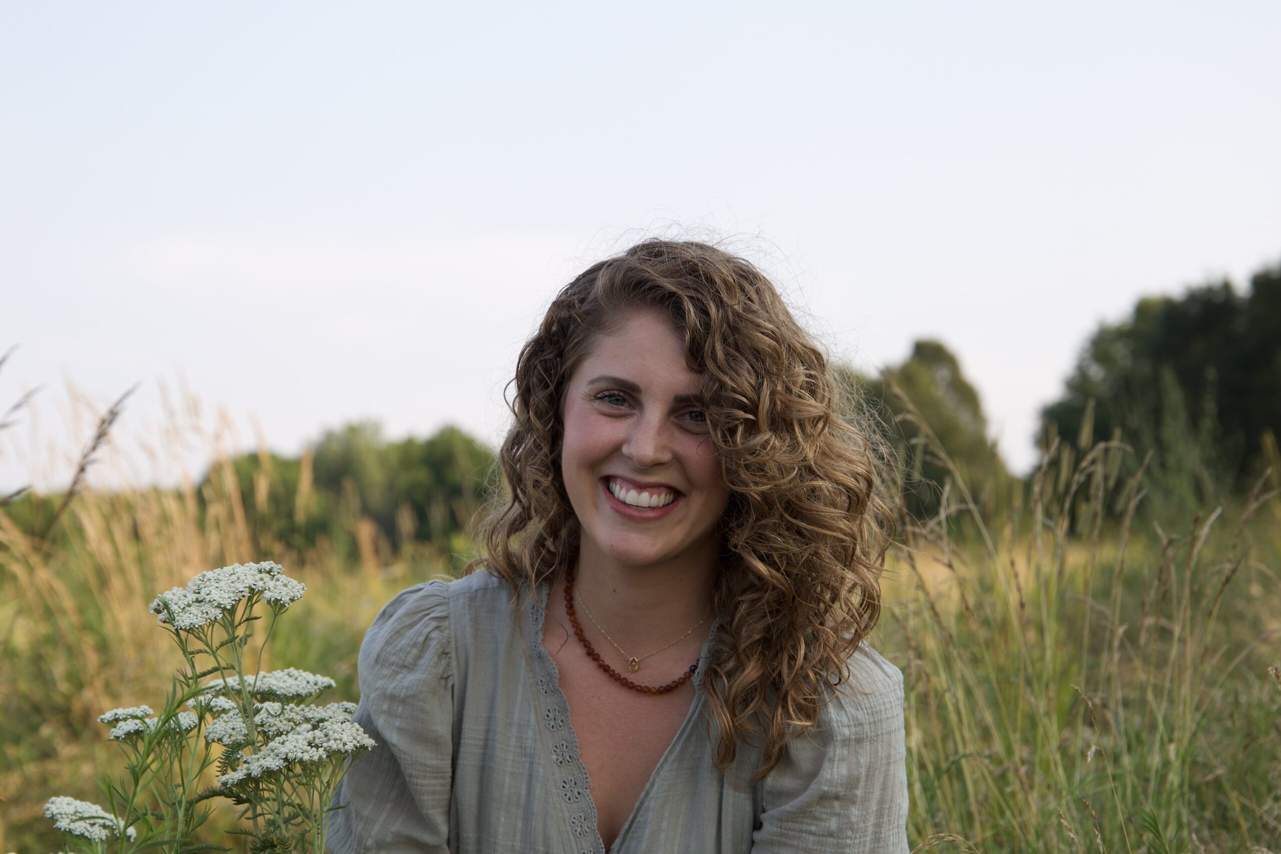 A woman with curly hair wearing a light gray blouse smiles while sitting in a grassy field dotted with wildflowers, perhaps taking a mindful break like many practitioners, with trees and a clear sky in the background.