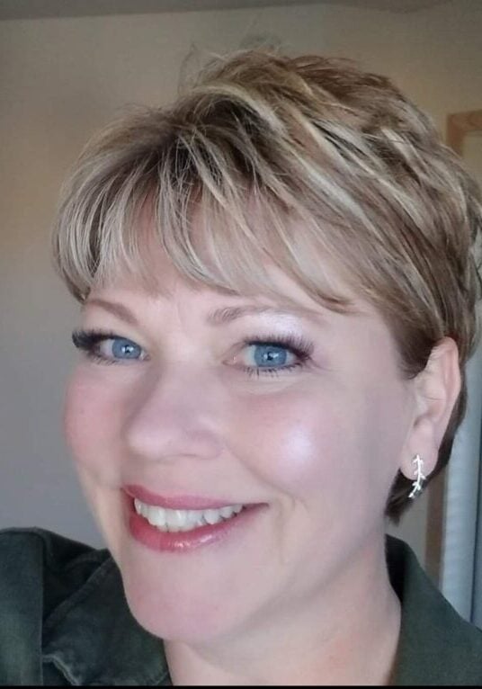 A woman with short, blonde hair and blue eyes smiles at the camera. She is wearing a dark green top, light makeup, and small dangling earrings. The bright room in the background suggests a welcoming space for practitioners.